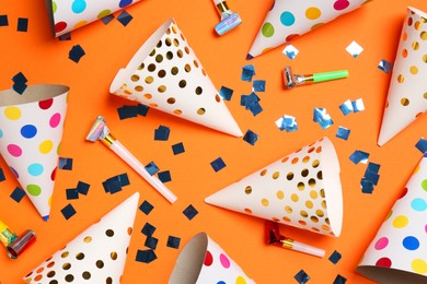 Shiny confetti, blowers and party hats on orange background, flat lay