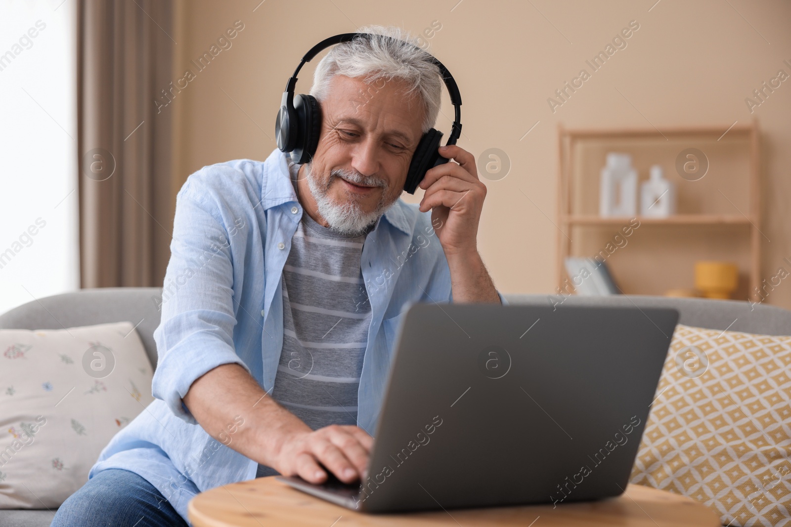 Photo of Senior man with headphones using laptop at wooden table indoors