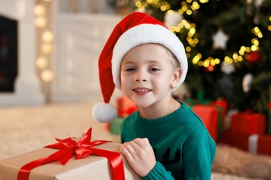 Photo of Cute little boy in Santa hat with Christmas gift indoors