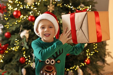 Photo of Cute little boy in Santa hat with Christmas gift indoors