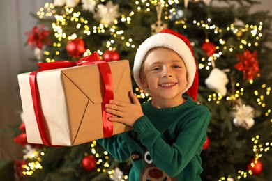 Photo of Cute little boy in Santa hat with Christmas gift indoors