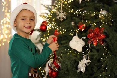 Photo of Cute little boy in Santa hat decorating Christmas tree at home