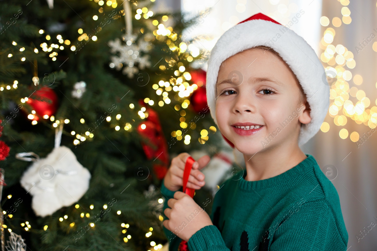 Photo of Cute little boy in Santa hat decorating Christmas tree at home