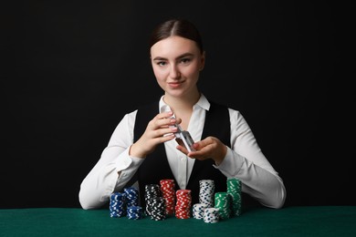 Professional croupier with casino chips and playing cards at gambling table on black background