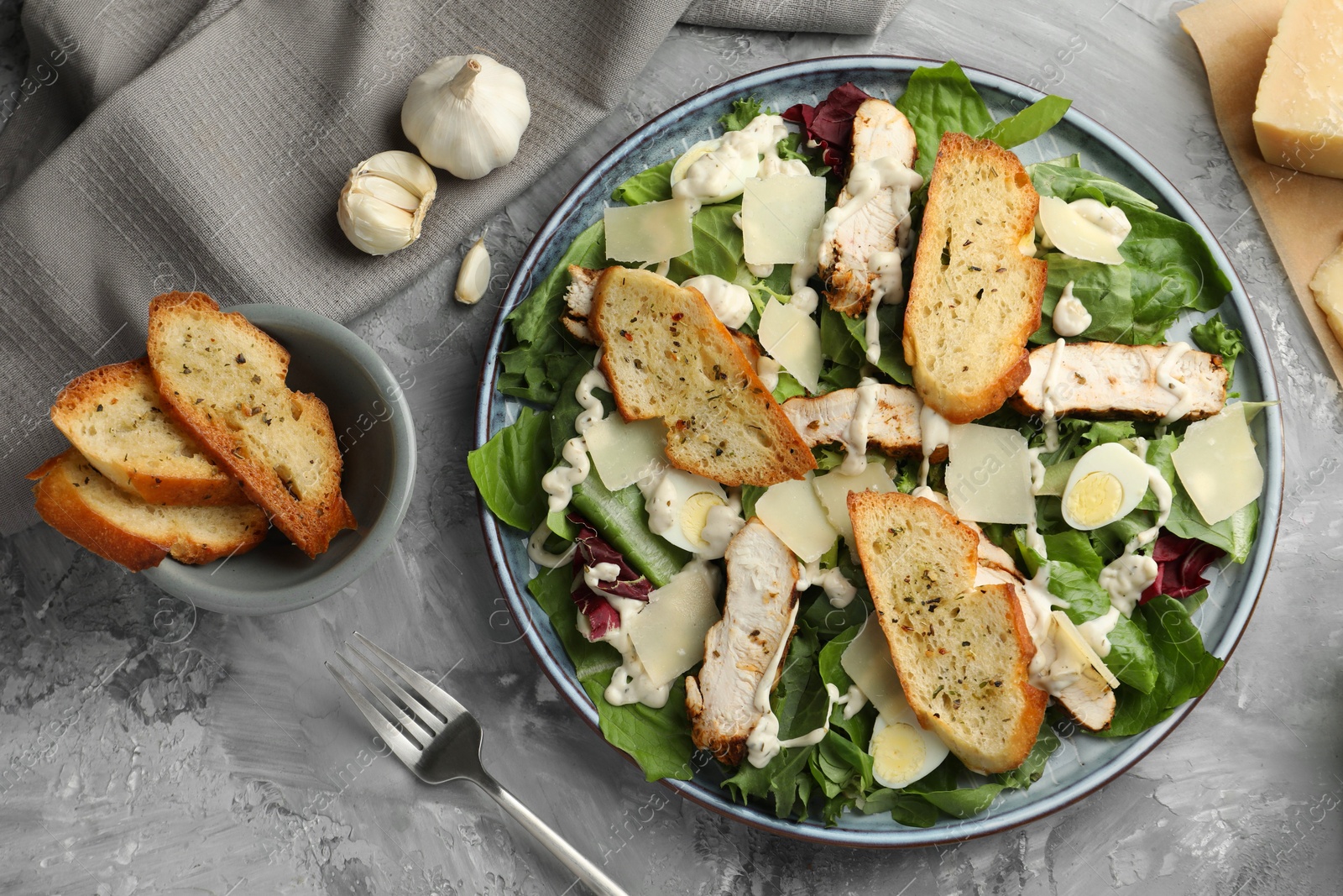 Photo of Tasty Caesar salad with chicken, ingredients and fork on gray textured table, flat lay