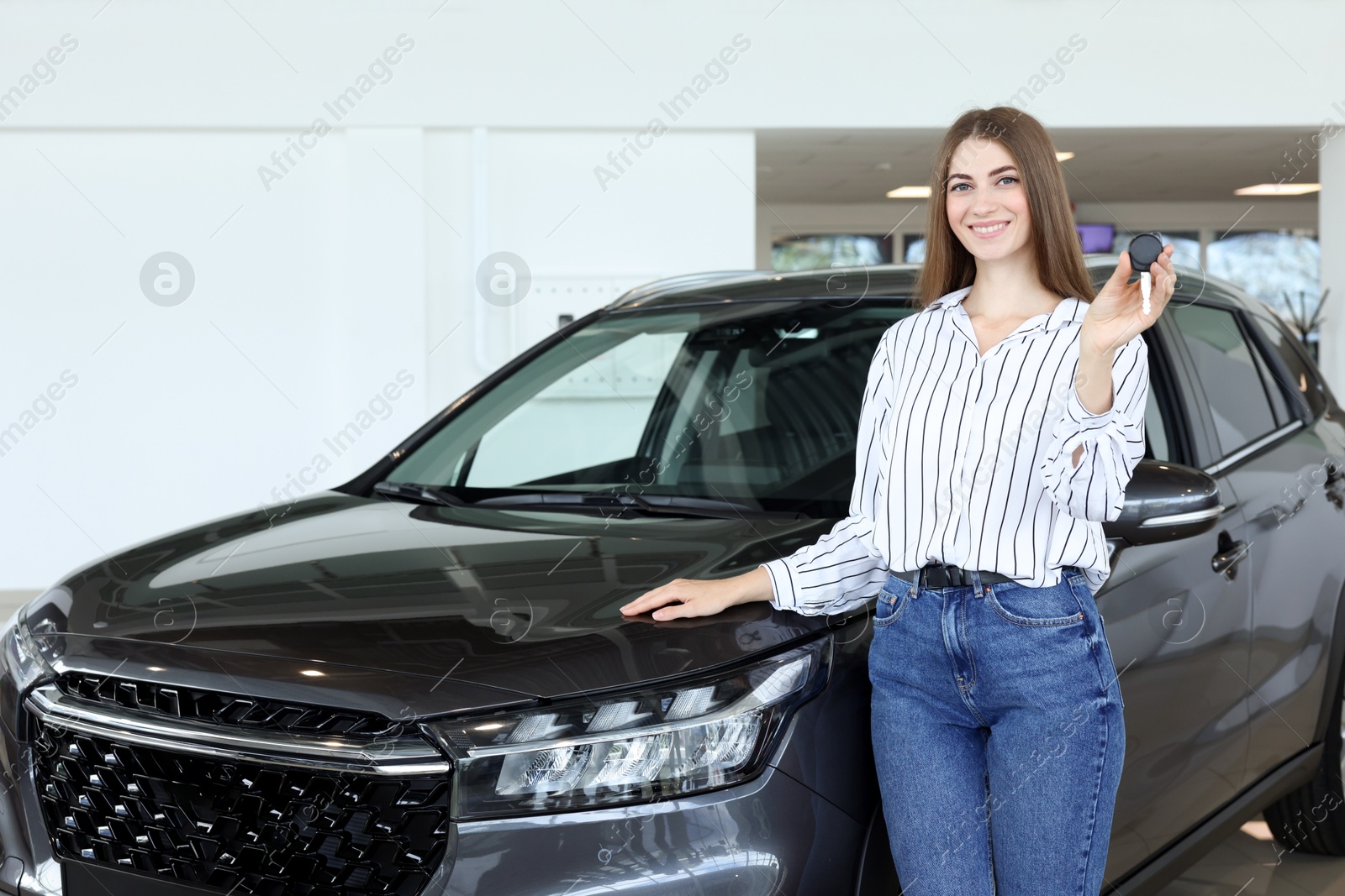 Photo of Happy young woman holding key near new black car in salon