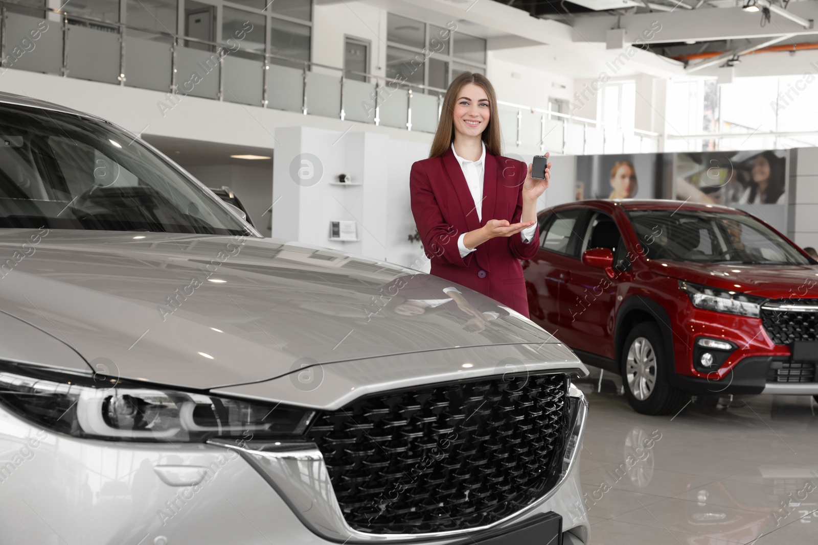 Photo of Happy saleswoman holding key near new silver car in salon