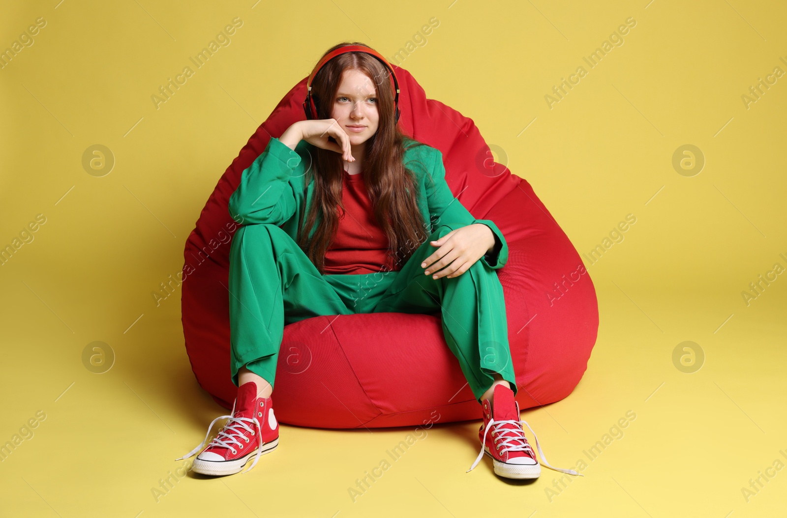 Photo of Teenage red-haired girl with headphones sitting on beanbag against golden background