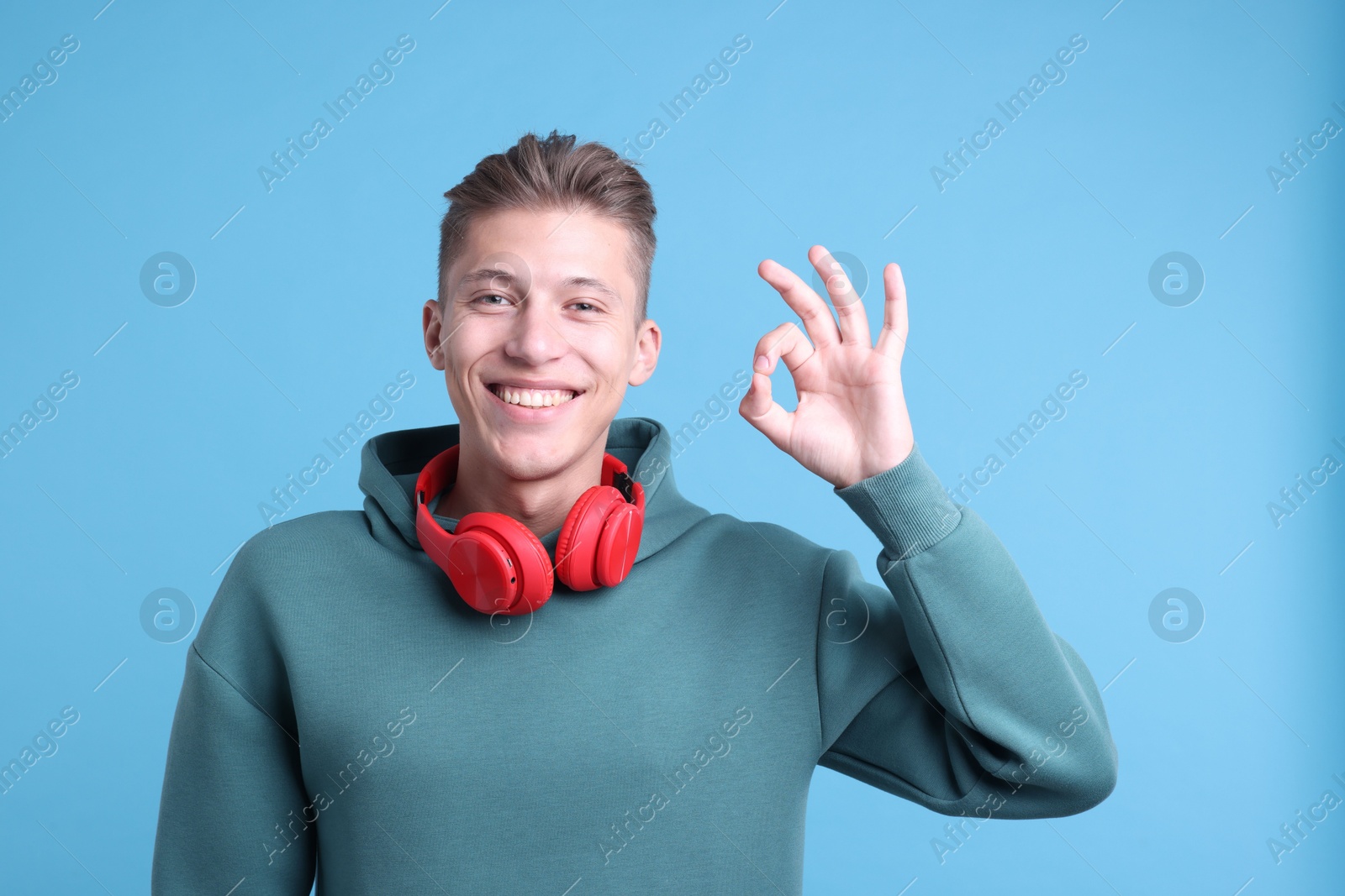 Photo of Happy young man with headphones showing OK gesture on light blue background