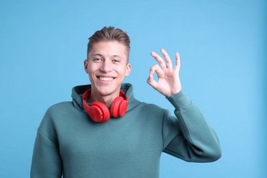 Happy young man with headphones showing OK gesture on light blue background