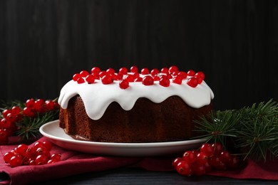 Photo of Tasty Christmas cake with red currants and fir branches on black wooden table against dark background, closeup