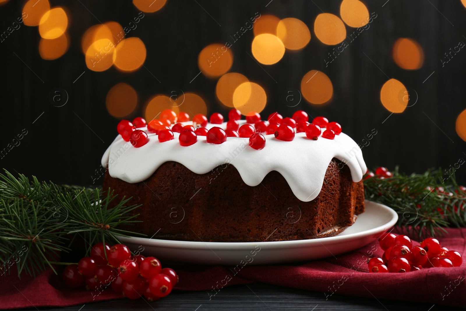 Photo of Tasty Christmas cake with red currants and fir branches on black wooden table against blurred lights, closeup