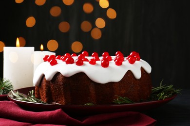 Photo of Tasty Christmas cake with red currants and rosemary on black wooden table against blurred lights, closeup