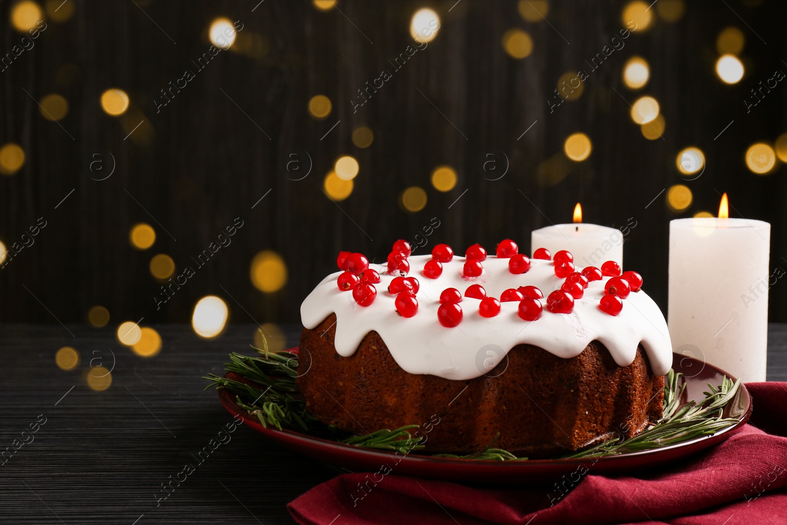 Photo of Tasty Christmas cake with red currants and rosemary on black wooden table against blurred lights, closeup