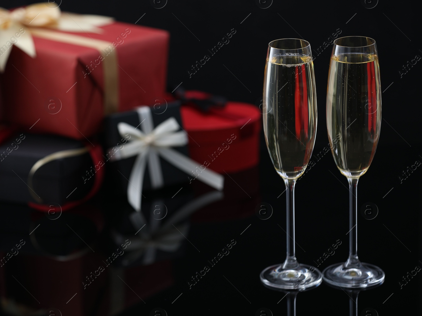 Photo of Delicious champagne in glasses and Christmas presents on black mirror surface, closeup. Space for text