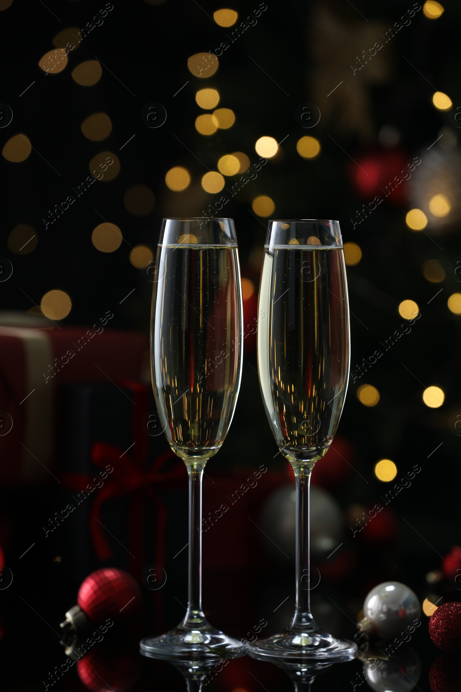 Photo of Delicious champagne in glasses and baubles on black mirror surface against blurred lights. Christmas celebration