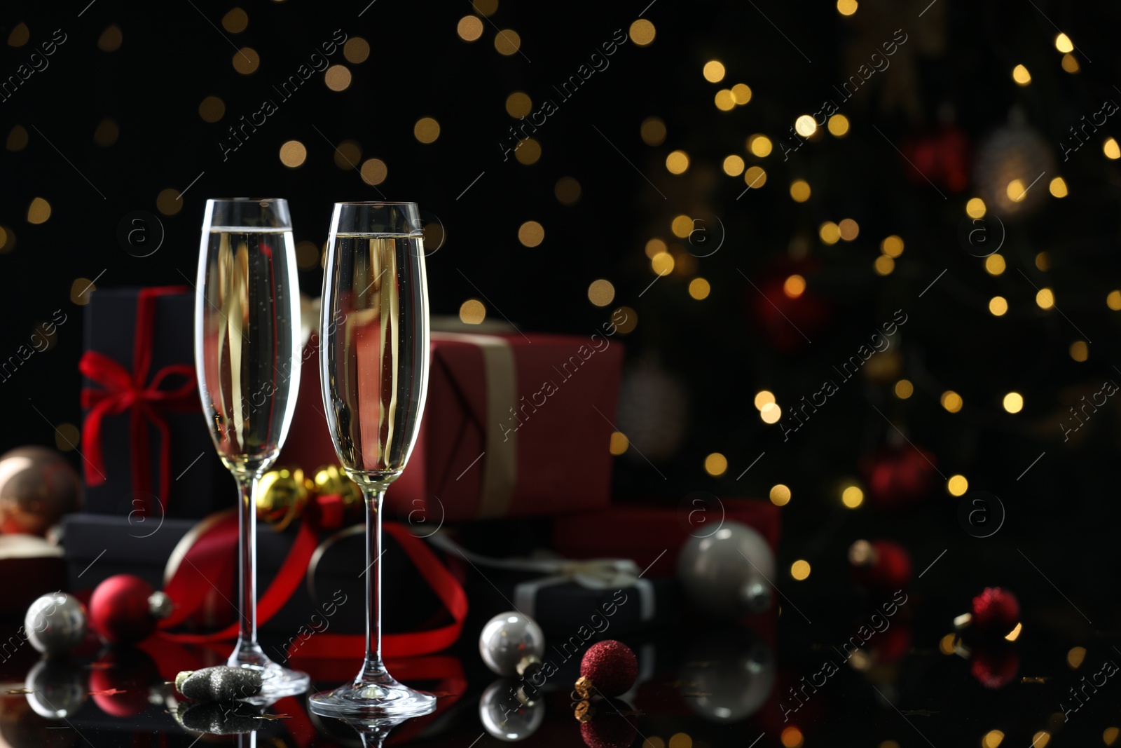 Photo of Delicious champagne in glasses and Christmas decor on black mirror surface against blurred lights. Space for text