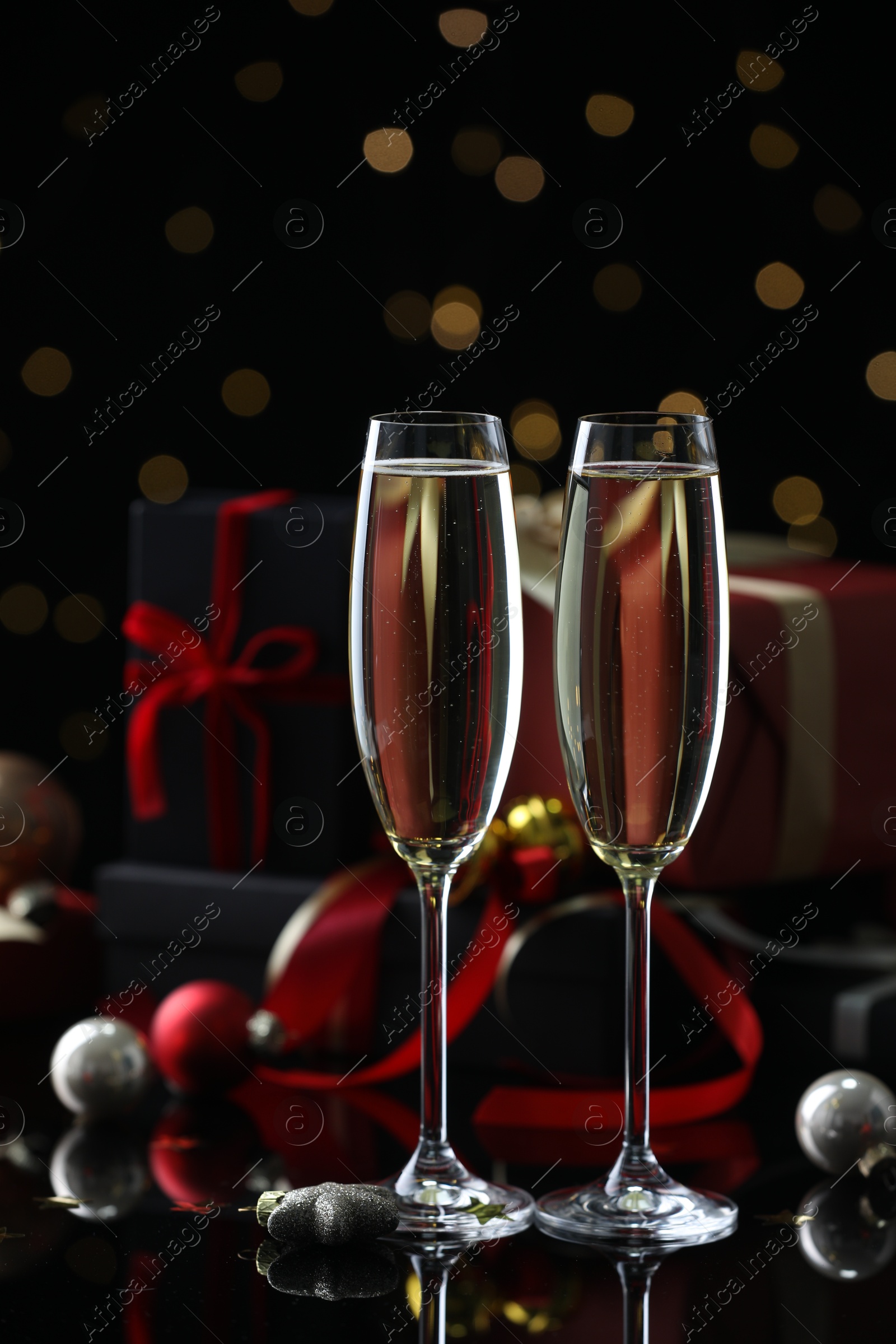 Photo of Delicious champagne in glasses and Christmas decor on black mirror surface against blurred lights