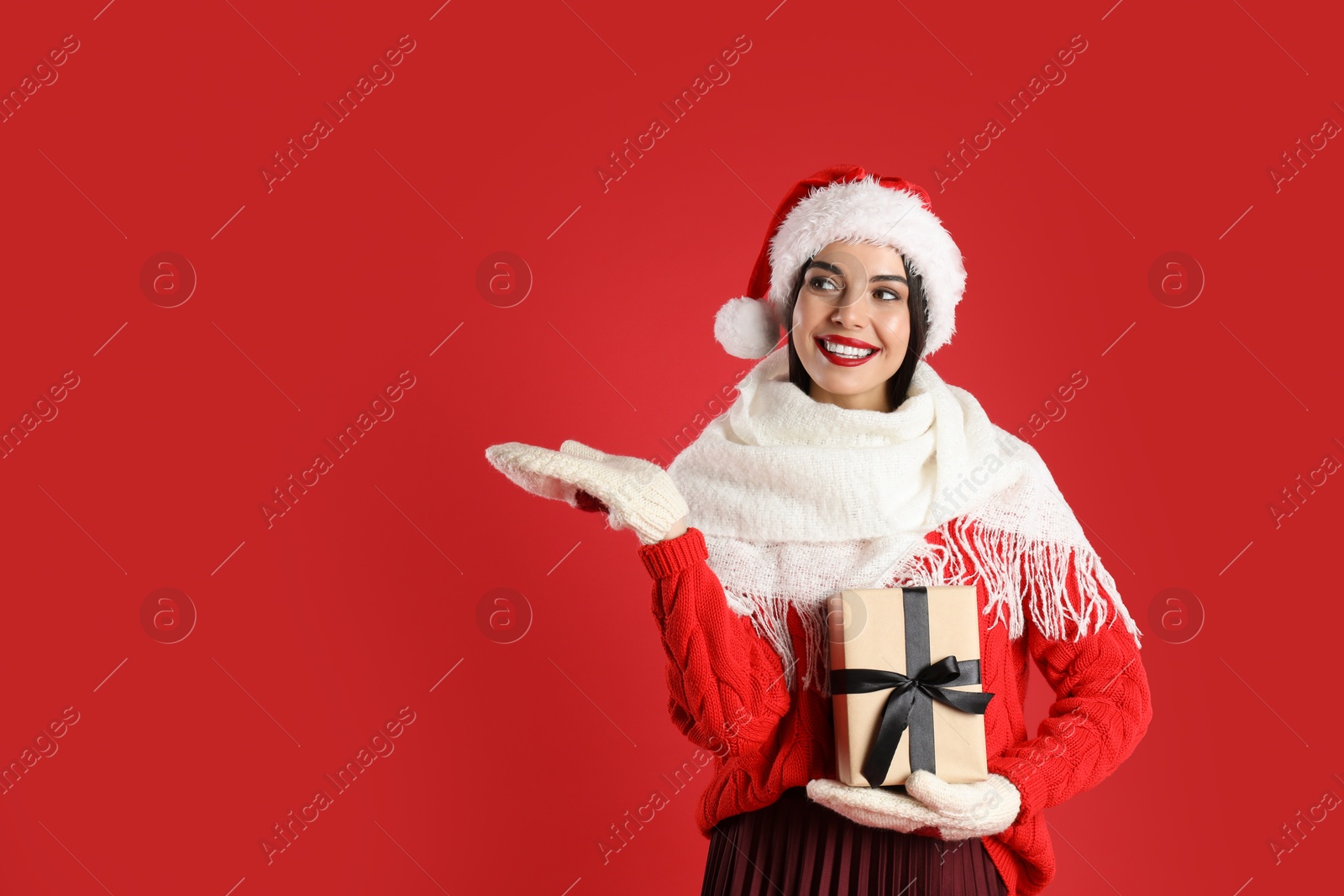 Photo of Woman in Santa hat, knitted mittens, scarf and sweater holding Christmas gift on red background, space for text