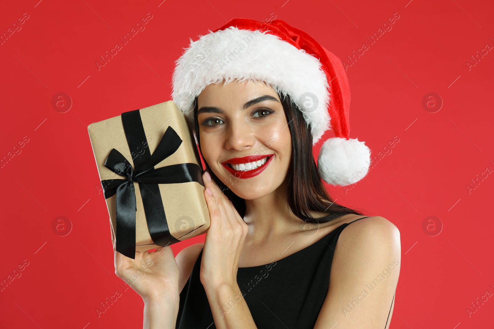 Photo of Woman in black dress and Santa hat holding Christmas gift on red background