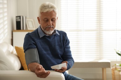 Senior man measuring blood pressure on sofa indoors