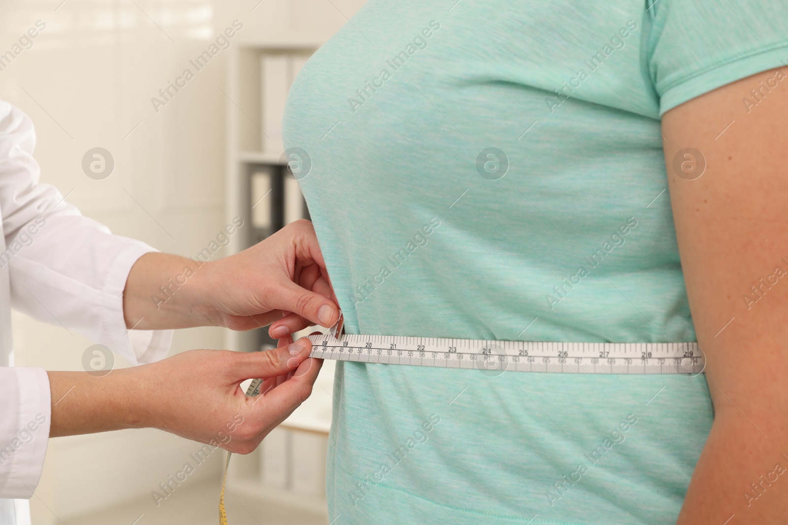 Photo of Nutritionist measuring overweight woman's waist with tape in hospital, closeup