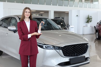 Photo of Happy saleswoman holding key near new silver car in salon