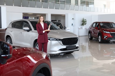 Photo of Happy saleswoman holding key near new silver car in salon