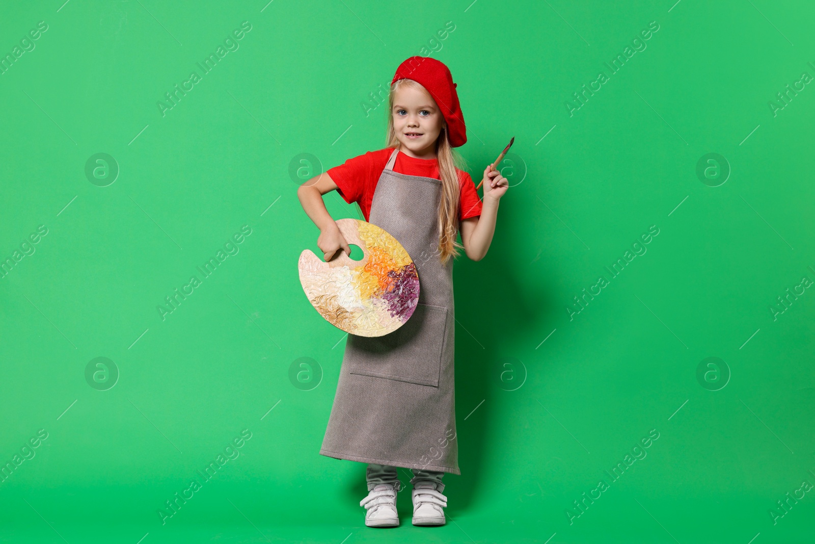 Photo of Little girl with brush and palette pretending to be artist on green background. Dreaming of future profession