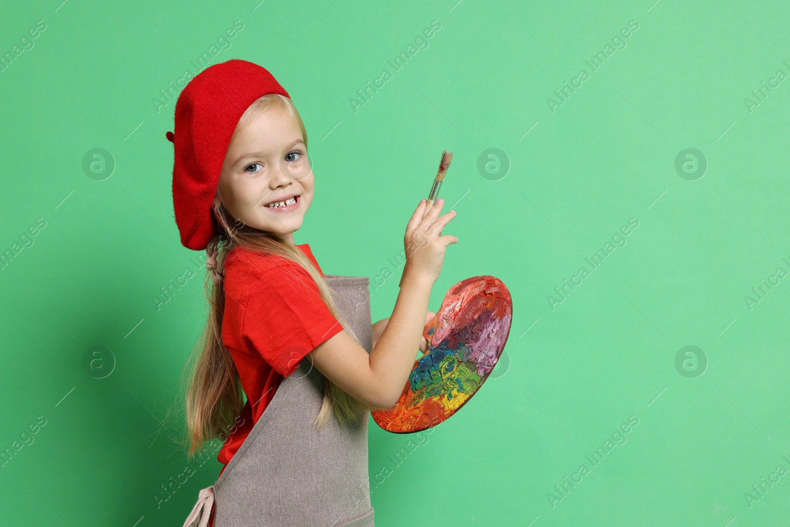 Photo of Little girl with brush and palette pretending to be artist on green background. Dreaming of future profession