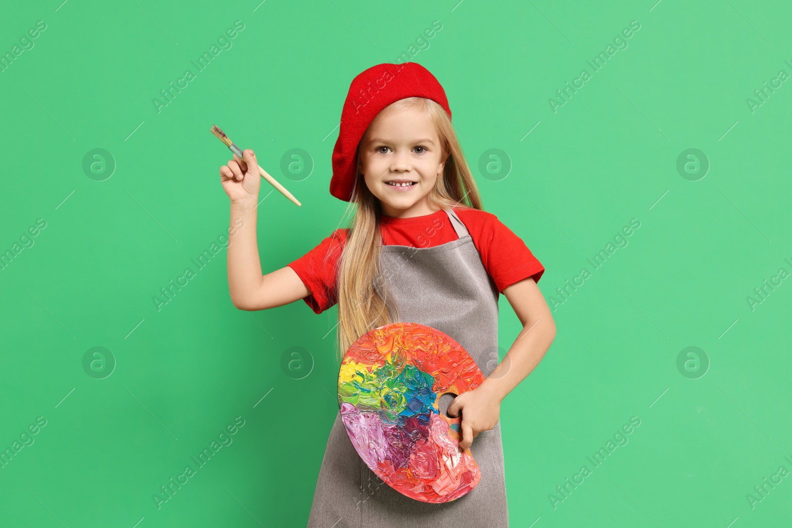 Photo of Little girl with brush and palette pretending to be artist on green background. Dreaming of future profession