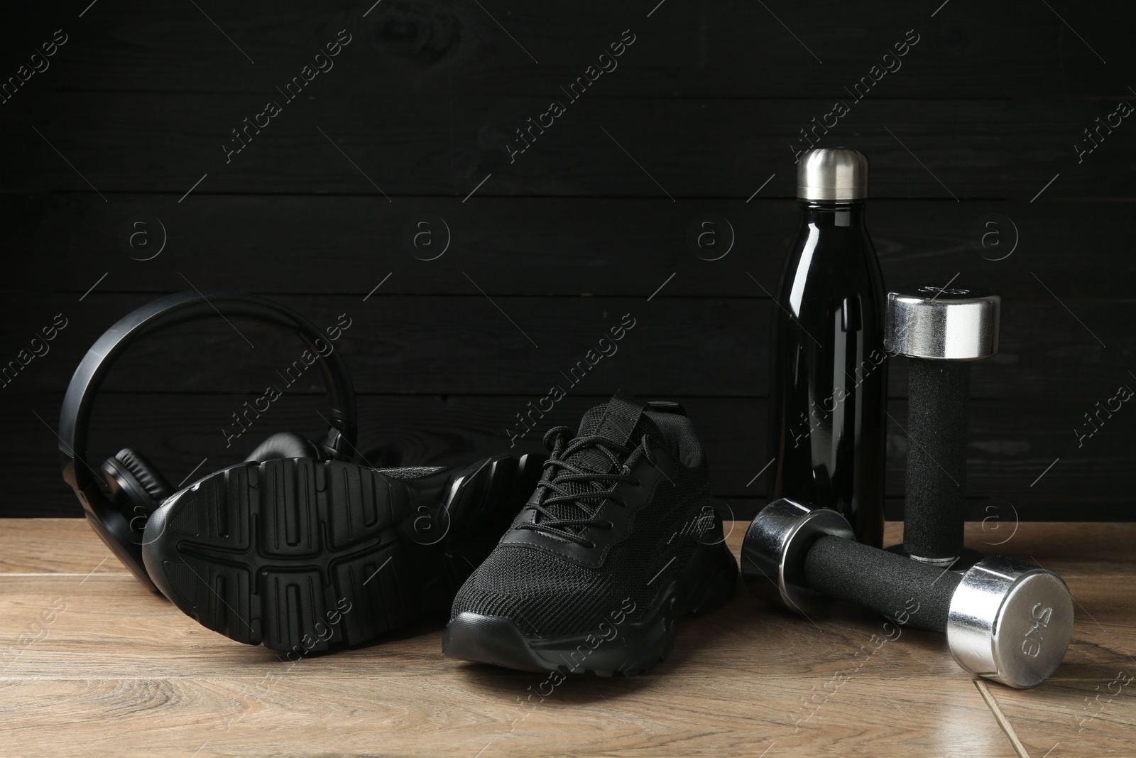 Photo of Pair of stylish black sneakers, dumbbells, thermo bottle and headphones on floor against dark wooden background