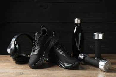 Photo of Pair of stylish black sneakers, dumbbells, thermo bottle and headphones on floor against dark wooden background