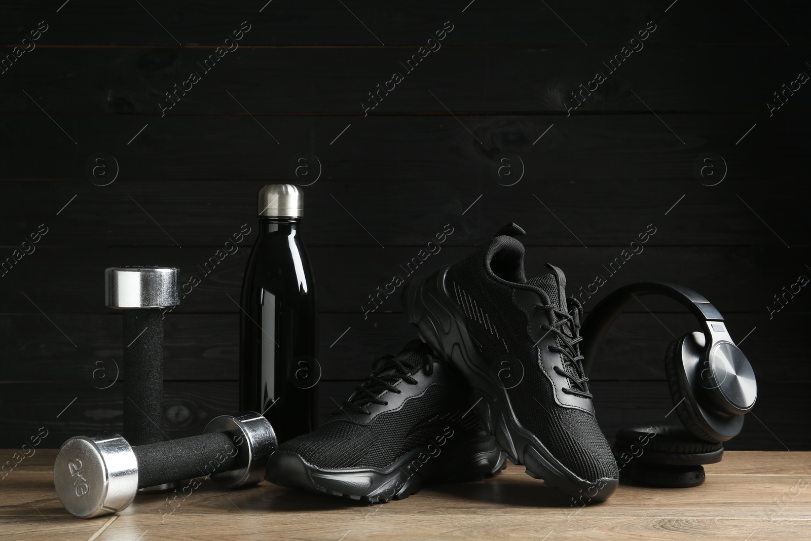 Photo of Pair of stylish black sneakers, dumbbells, thermo bottle and headphones on floor against dark wooden background