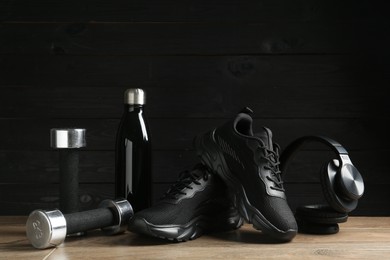 Photo of Pair of stylish black sneakers, dumbbells, thermo bottle and headphones on floor against dark wooden background