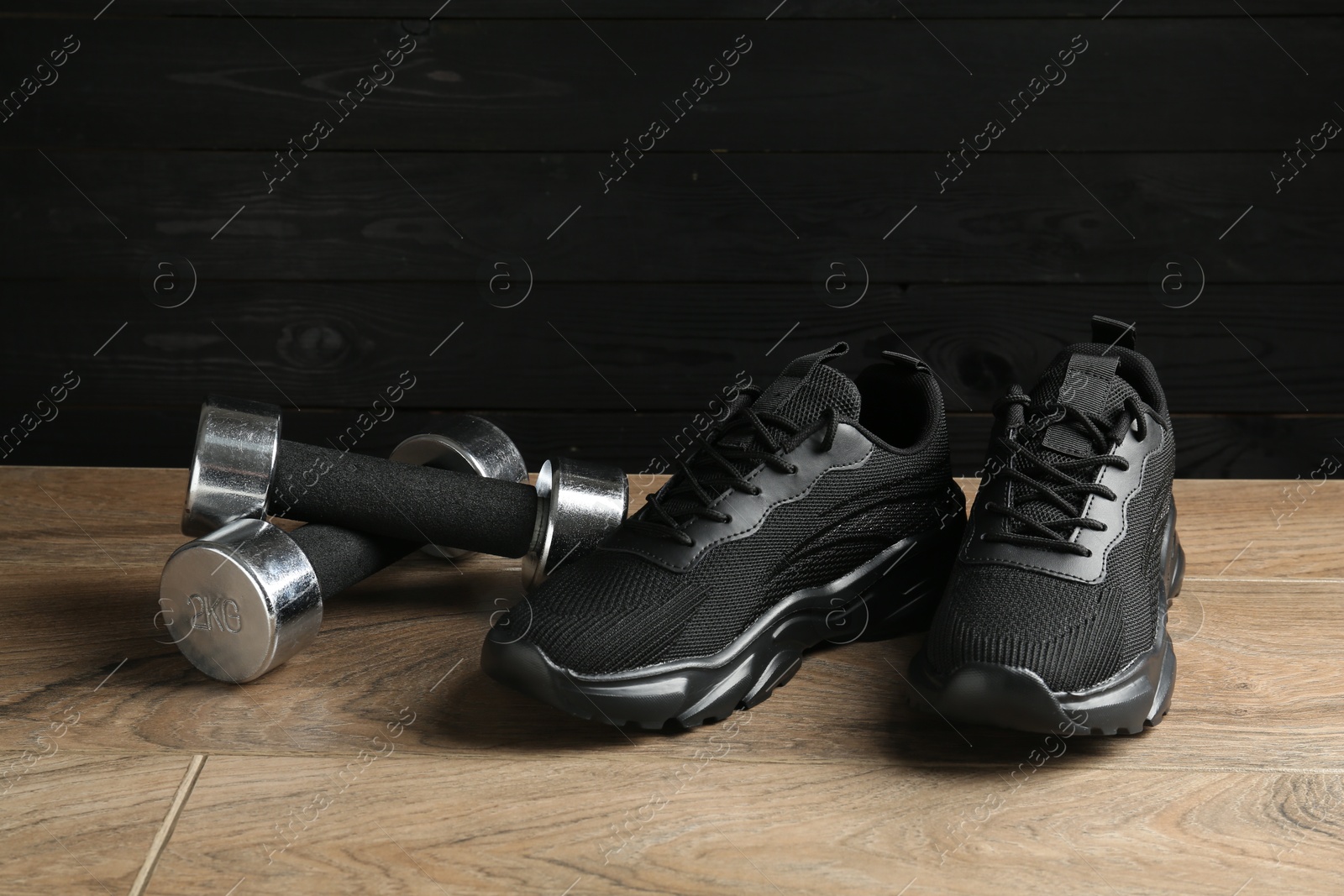 Photo of Pair of stylish black sneakers and dumbbells on floor against dark wooden background