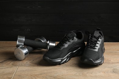 Photo of Pair of stylish black sneakers and dumbbells on floor against dark wooden background