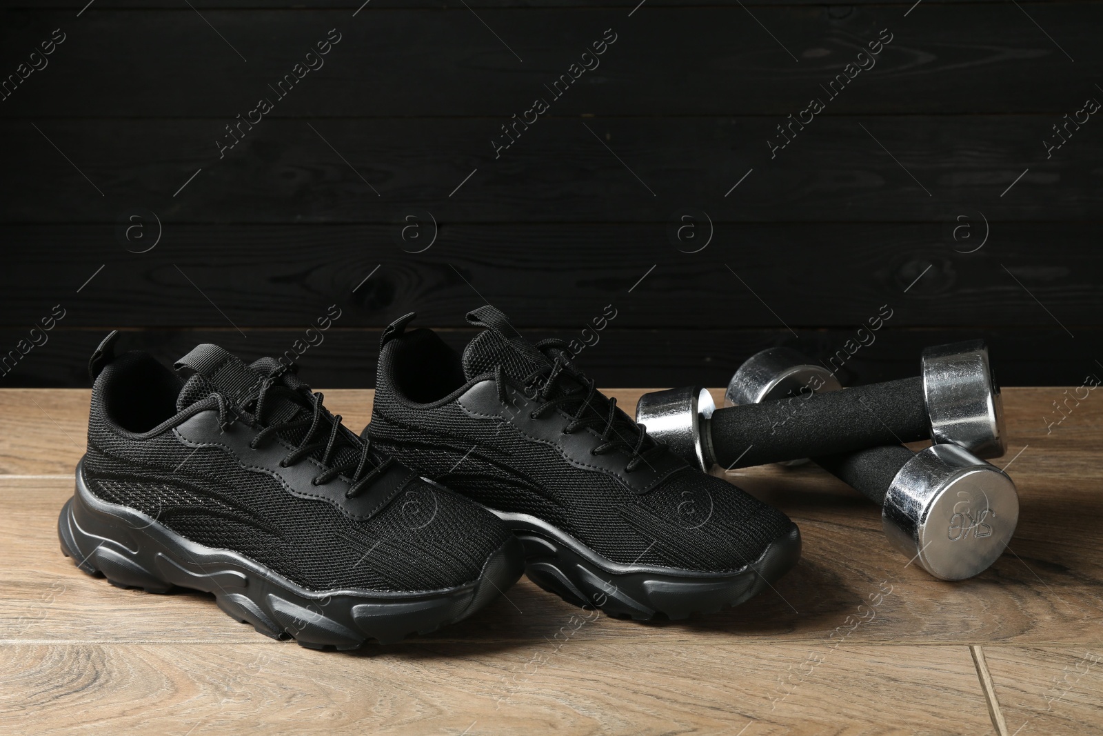 Photo of Pair of stylish black sneakers and dumbbells on floor against dark wooden background