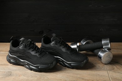 Photo of Pair of stylish black sneakers and dumbbells on floor against dark wooden background