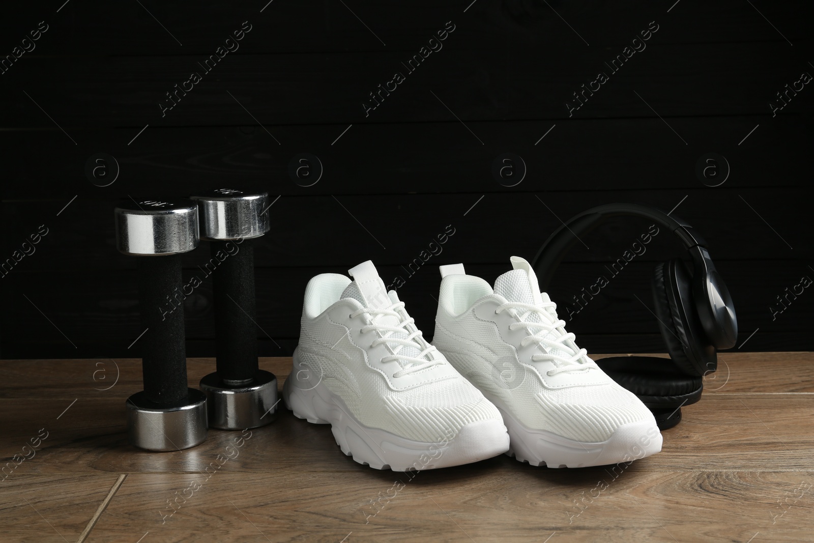 Photo of Pair of stylish white sneakers, dumbbells and headphones on wooden floor against dark background