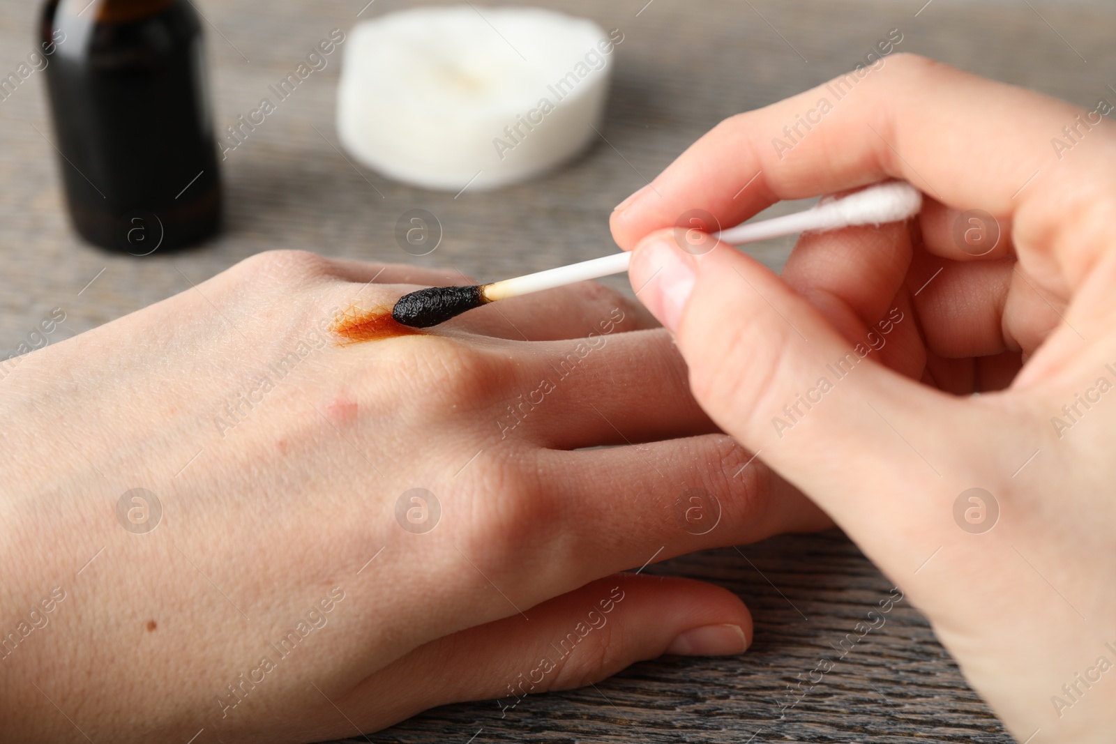 Photo of Woman applying topical iodine with cotton swab on hand at wooden table, closeup