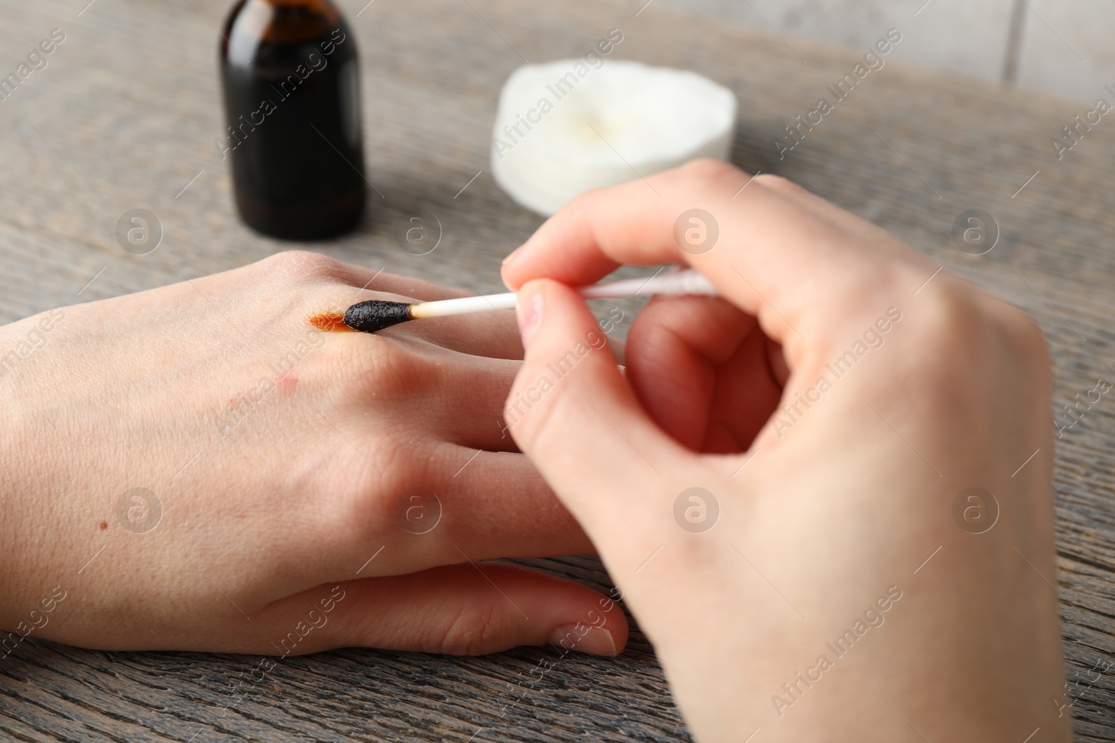 Photo of Woman applying topical iodine with cotton swab on hand at wooden table, closeup