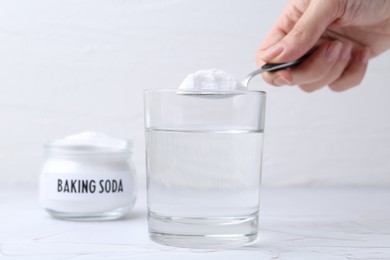 Photo of Woman adding baking soda to glass of water at white textured table, closeup