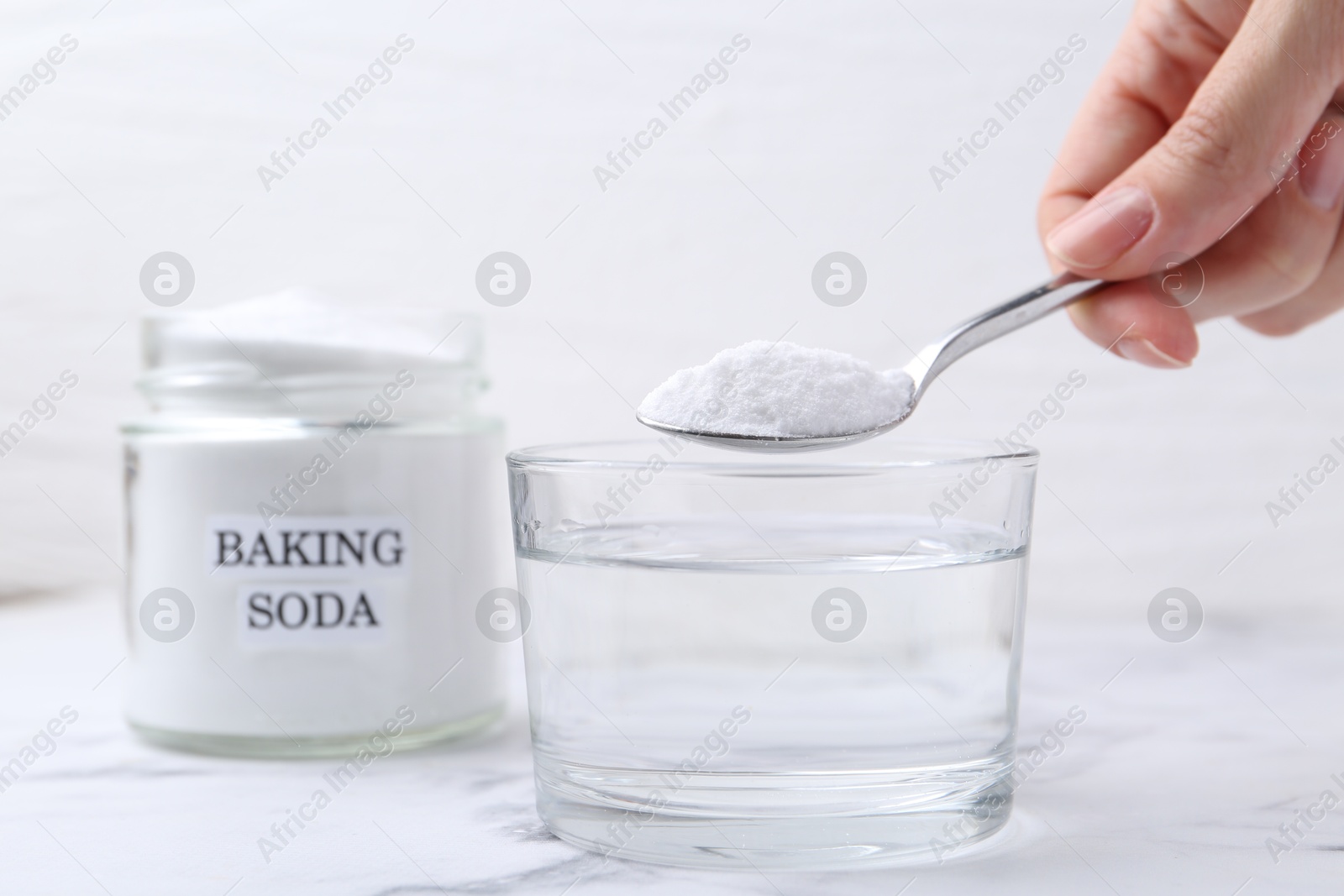 Photo of Woman adding baking soda to glass of water at white marble table, closeup