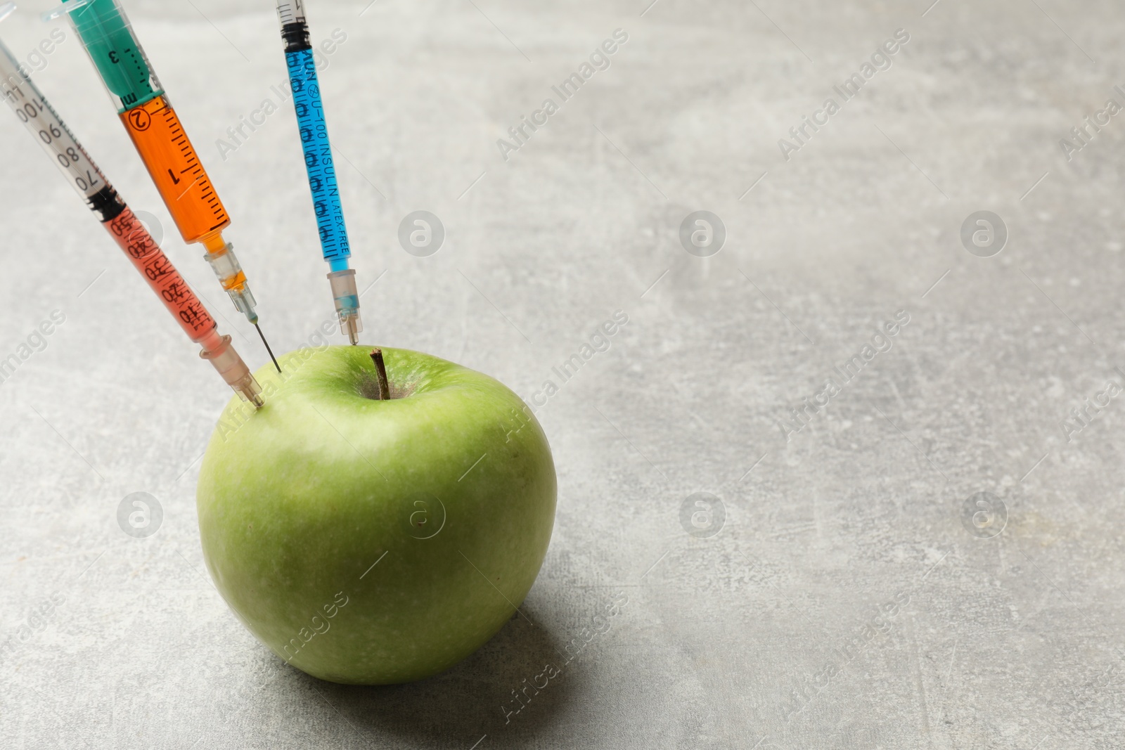 Photo of GMO concept. Green apple with different syringes on grey textured table, closeup. Space for text