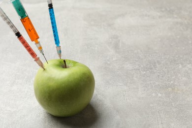 Photo of GMO concept. Green apple with different syringes on grey textured table, closeup. Space for text