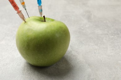 Photo of GMO concept. Green apple with different syringes on grey textured table, closeup. Space for text