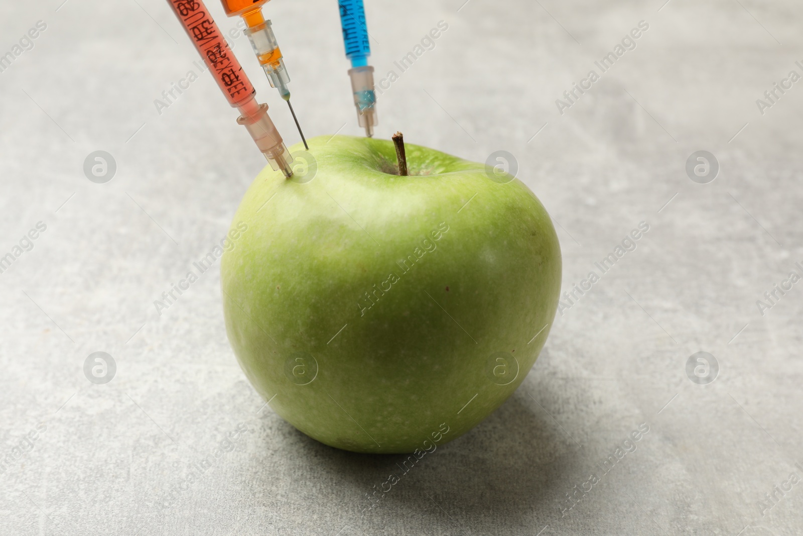 Photo of GMO concept. Green apple with different syringes on grey textured table, closeup