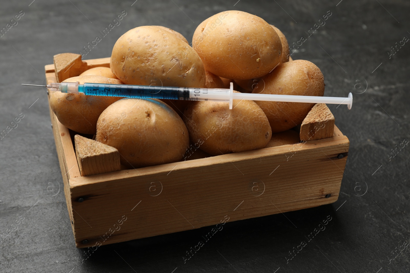 Photo of GMO concept. Potatoes and syringe with liquid in crate on dark textured table, closeup