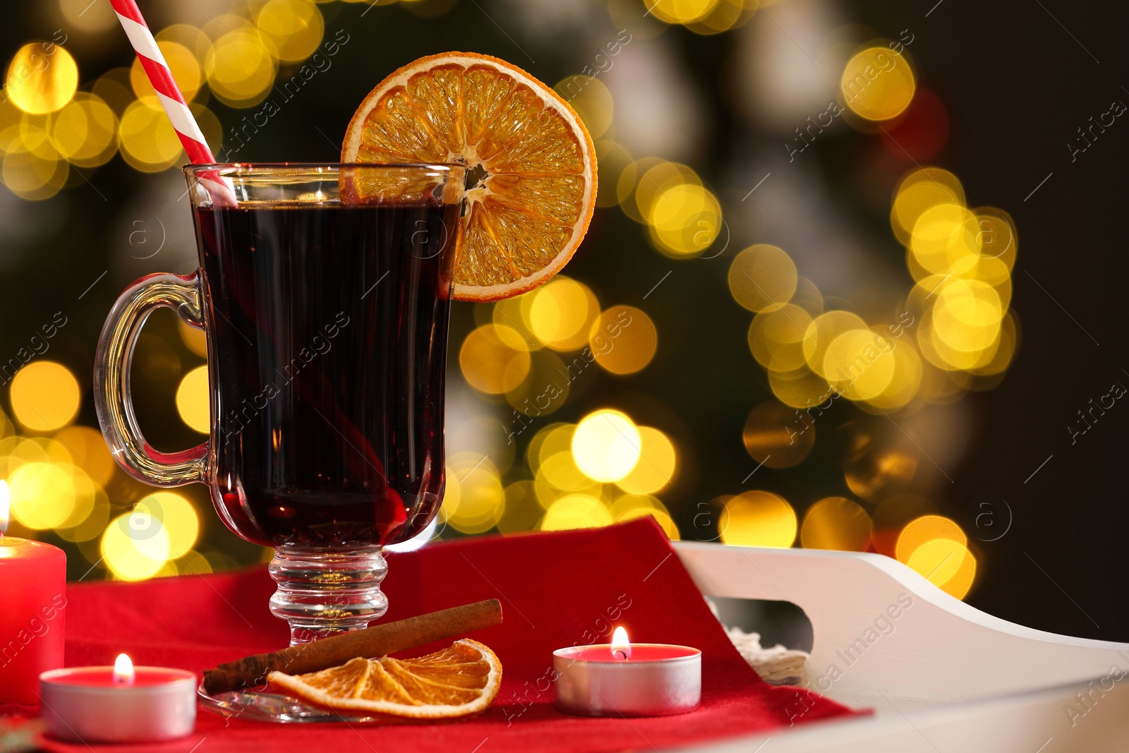 Photo of Aromatic mulled wine in glass cup and burning candles on tray against blurred Christmas lights, space for text. Bokeh effect